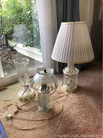 Three lead crystal lamps of different sizes placed on a carpet near a window showing bases and one with a white fabric shade.