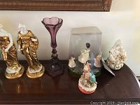 Photo showing various porcelain figurines including gold-accented draped women, a purple glass vase, small figurines on and under a dome, and a white and gold seated figure.