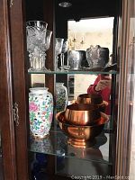 Glass shelving with etched glass wine glasses on top shelf, two decorative ceramic vases with floral patterns on bottom shelf, and three copper bowls stacked together.