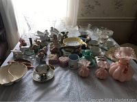 Wide view showing assortment of clear cut glass bowls, various porcelain animal figurines, teacups, and other ceramic decorative items arranged on a table with a white lace tablecloth.