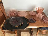 Photo showing an assortment of pink Depression glass pieces and black tole trays arranged on a wooden table, including a creamer pitcher, covered jar, several plates, and multiple trays.