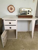 Full view of white wooden sewing desk with vintage Kenmore sewing machine on top, wall clock in background.