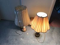 Three vintage lamps on floor, two with beige pleated shades upright and one with shade wrapped/covered, cords visible, one lamp lit