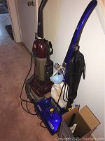 Side view of two upright vacuums Hoover and Eureka standing against a wall showing power cords and handles.
