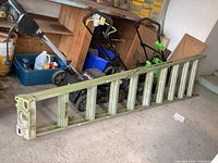 Photo of a 10-foot aluminum ladder lying flat on a concrete floor in a garage or storage area, surrounded by other tools and equipment.