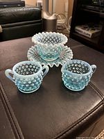 Four pieces of blue opalescent hobnail glassware including cream pitcher, sugar bowl, small bowl, and matching ruffled-edge saucer.