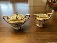 Front view of hand painted Nippon sugar bowl with lid and matching cream pitcher on wooden table.
