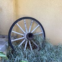 Front view of one large vintage wagon wheel leaning against a yellow wall, resting on ground with vegetation at base