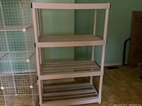 Front view of beige/light gray plastic four-tier utility shelving unit standing in a room with green walls and brown carpet.