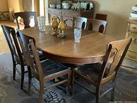 Dining table with leaf extended, surrounded by six oak chairs with black leather seats. Table shows medium brown wood finish with round pedestal base.