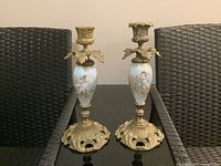 Pair of two French candlestick holders with porcelain bodies and brass bases, placed on black reflective surface between wicker chairs.