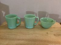 Three Fire King Jadeite pieces, two mugs and one cup, shown side by side on a wooden table against a plain background.