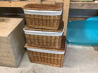 Three woven wicker utility baskets stacked, showing natural brown wicker and white cotton liners. Large basket measures 23 inches long by 14 inches high.