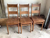 Front view showing the three solid wood chairs side by side against a concrete floor and wall, highlighting their shape and construction.