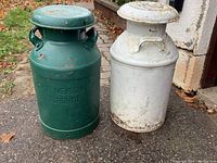 Front view of two metal milk cans, one green with embossing, one plain white, both showing wear and rust.
