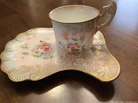 Photo showing the porcelain cup with an ornate handle and a matching scalloped saucer with gold accents and floral decoration.