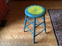 Full view of the blue painted wooden stool standing on parquet floor, showing its four legs and round seat.