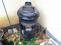 Full view of the vintage Martin stove outdoors next to a wall and plants.