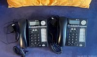 Two vintage black AT&T 2 line desk phones placed side by side with handsets and visible cords, showing digital display, numeric keypad and function keys.