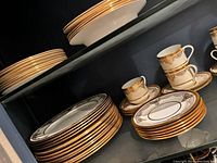 Shelved collection showing stacked Aynsley Trafalgar bone china plates and cups with gold decorative rims.