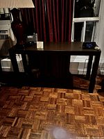 Photo of a rectangular brown Parsons table with square legs placed against a wall with red curtains and windows in the background, showing the top surface and leg details.