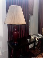 Photo of a classic vintage burgundy table lamp with a tall pleated beige lampshade placed on a black table against burgundy curtains.