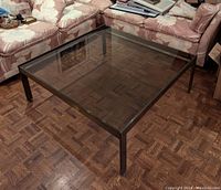 Full view of square coffee table showing brass frame and smoked glass top, placed on parquet flooring.