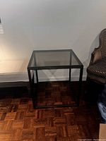 Square coffee table with metal frame and smoked glass top positioned on a parquet floor, flanked by chairs.