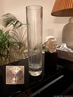 Photo of a tall clear glass vase with etched vertical patterns on a black surface, next to a lamp and decorative objects.