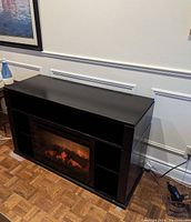 Black electric fireplace with faux flames lit inside and side shelves, shown against a white wall with molding and parquet floor.