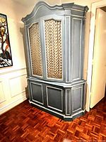 Vintage French Provincial armoire with detailed molding, lattice work doors with curtains, painted finish, placed against a wall in a room.