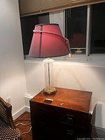 Photo of a vintage slimline glass table lamp with a pink fabric shade resting on a wooden side table. The lamp is turned on, showing the light through the shade. The base is clear textured glass with a round brass-colored base.