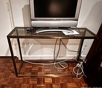 Photo showing the table TV stand with a flat screen TV on top, power cables and remote controls visible on the wooden floor below the stand.