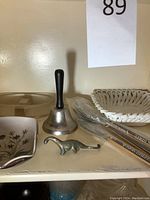 Top shelf view showing clear glass container, white floral bowl, metal bell, metal dinosaur, skewers and woven basket