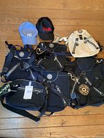 All items arranged on wooden floor showing black and beige Kipling bags and two caps