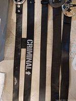 Seven black belts laid on tile floor showing various buckle designs including round silver, studded rectangular, O-ring, plain, printed “CRIMINAL”, and cross-design buckles.