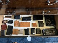 Overhead photo of various leather wallets and a luggage tag displayed inside a drawer, showing colors ranging primarily from black to brown with one red wallet. Various sizes and styles including small, bi-fold, zippered, and embossed leather wallets.
