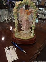 Angel figurine in pink dress holding a candle in floral arch mounted on wooden base