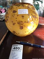 Full view of spherical yellow art glass vase with gold flecks on wooden surface, includes note card and pen for scale