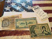 Three military-related paper currency notes laid on a textured cloth background. Included are a British Military Authority note, a 1 schilling Allied Military Authority note, and a 5 dinar note from Algeria with Arabic and English script.