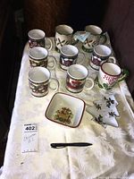Photo showing six Christmas-themed mugs, two creamers, one Christmas tree shaped plate, and one square small plate on a cream tablecloth.