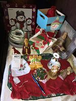 Photo of assorted Christmas-themed decorations including framed snowman art, wooden boxes, ceramic plates, cloth bag, placemats, small towels, figurines, and other decorative items on red fabric surface.