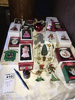 Full overview of many boxed Hallmark ornaments and loose items arranged on tablecloth, including red ball, figurines and angel ornaments.