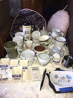Photo showing collection of assorted cups, mugs, creamers, and small ceramic containers with labels on a table.