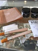 Image showing gold leaf sheets, two jars labeled varnish and gilding adhesive, two flat brushes, disposable gloves, and small metal craft accessories on a glass surface.