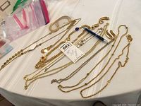 Top-down image of nine assorted gold tone necklaces laid flat on a white surface with a blue pen for scale and a plastic bag in background.