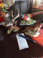 Photo showing three porcelain bird figurines placed on wooden surface with a pen and handwritten lot tag "Pretty Birds #4".