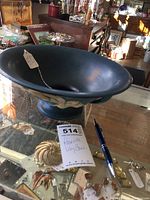 Blue Roseville lily bowl with yellow raised flower decoration and pedestal base, placed on glass display with assorted jewelry around it, alongside a pen and auction lot tag.
