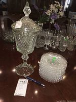 Tall clear glass compote with intricate diamond cut pattern and a lid with pointed finial, alongside nine clear glass appetizer plates with hobnail pattern stacked next to it.