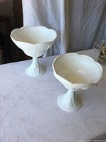 Two white milk glass compotes with grape and leaf relief designs, different sizes, placed on table near curtain backdrop.